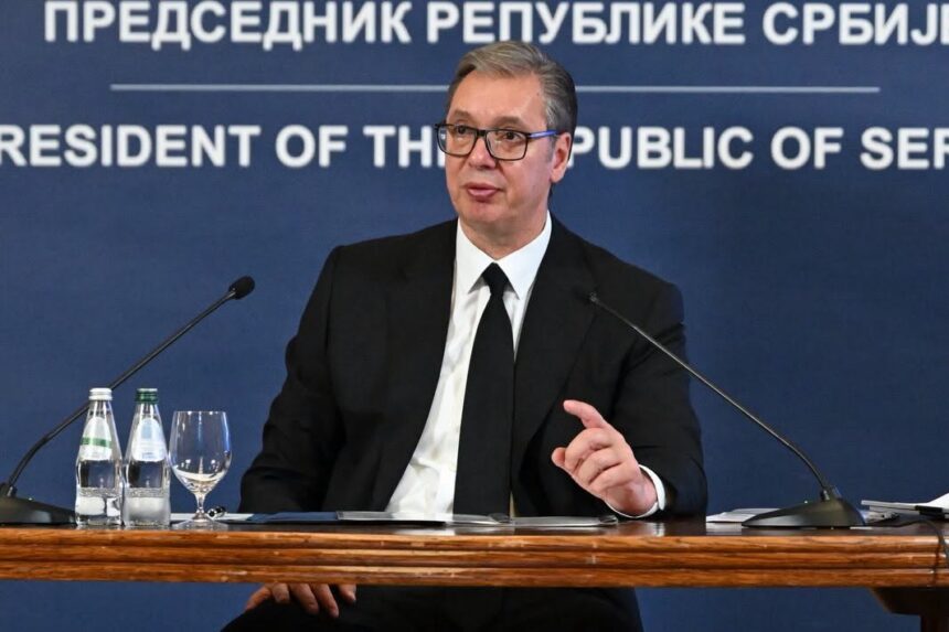 Man in a suit and glasses speaks at a press conference, seated behind a wooden table with microphones and water bottles. Blue backdrop reads 'President of the Republic of Serbia'.