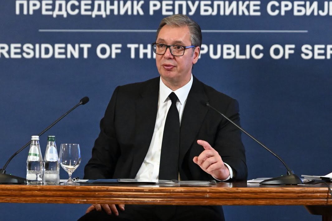 Man in a suit and glasses speaks at a press conference, seated behind a wooden table with microphones and water bottles. Blue backdrop reads 'President of the Republic of Serbia'.