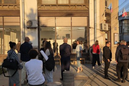 Group of people gathered outside a beige building with ornate decorative windows and a glass storefront, lit by warm afternoon sun.