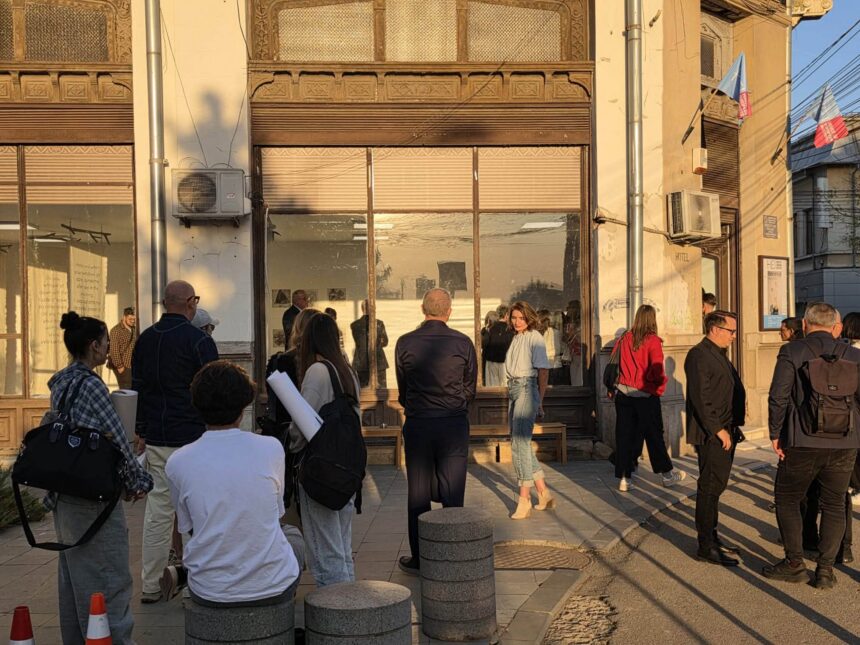 Group of people gathered outside a beige building with ornate decorative windows and a glass storefront, lit by warm afternoon sun.