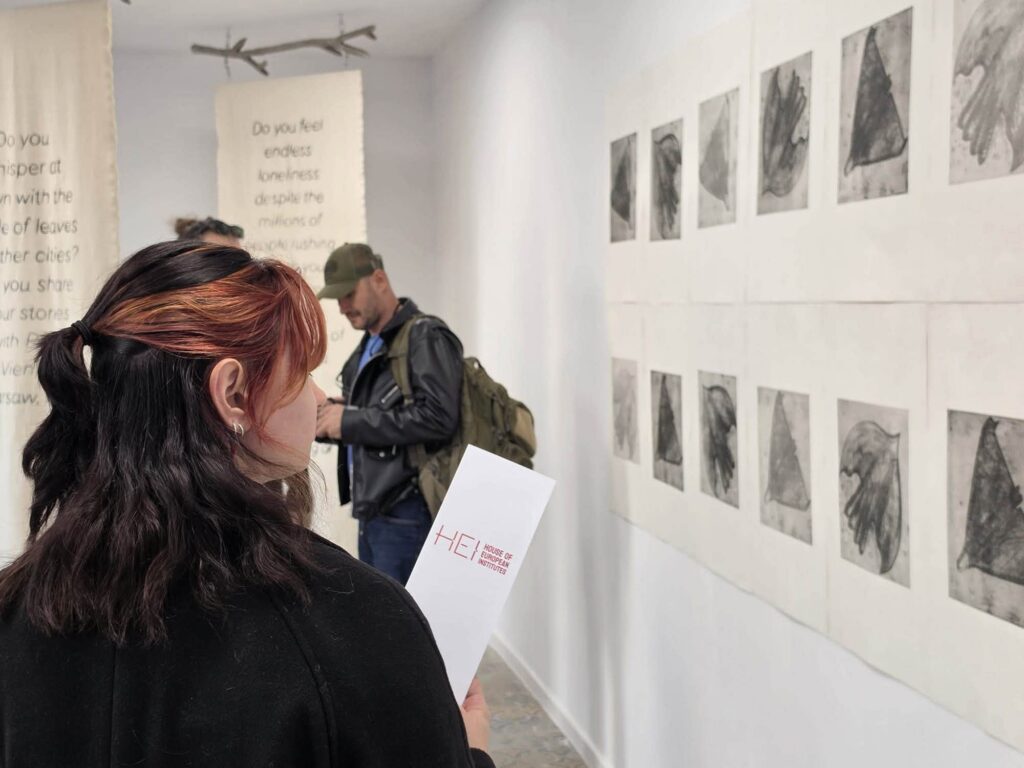 Woman with burgundy-streaked hair stands in the foreground, looking at a row of grayscale art prints along a white gallery wall as a man with a backpack browses nearby.