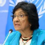 Woman speaking at a UN press briefing, wearing a turquoise blazer and beaded necklace, microphone in front.