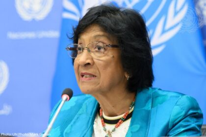 Woman speaking at a UN press briefing, wearing a turquoise blazer and beaded necklace, microphone in front.