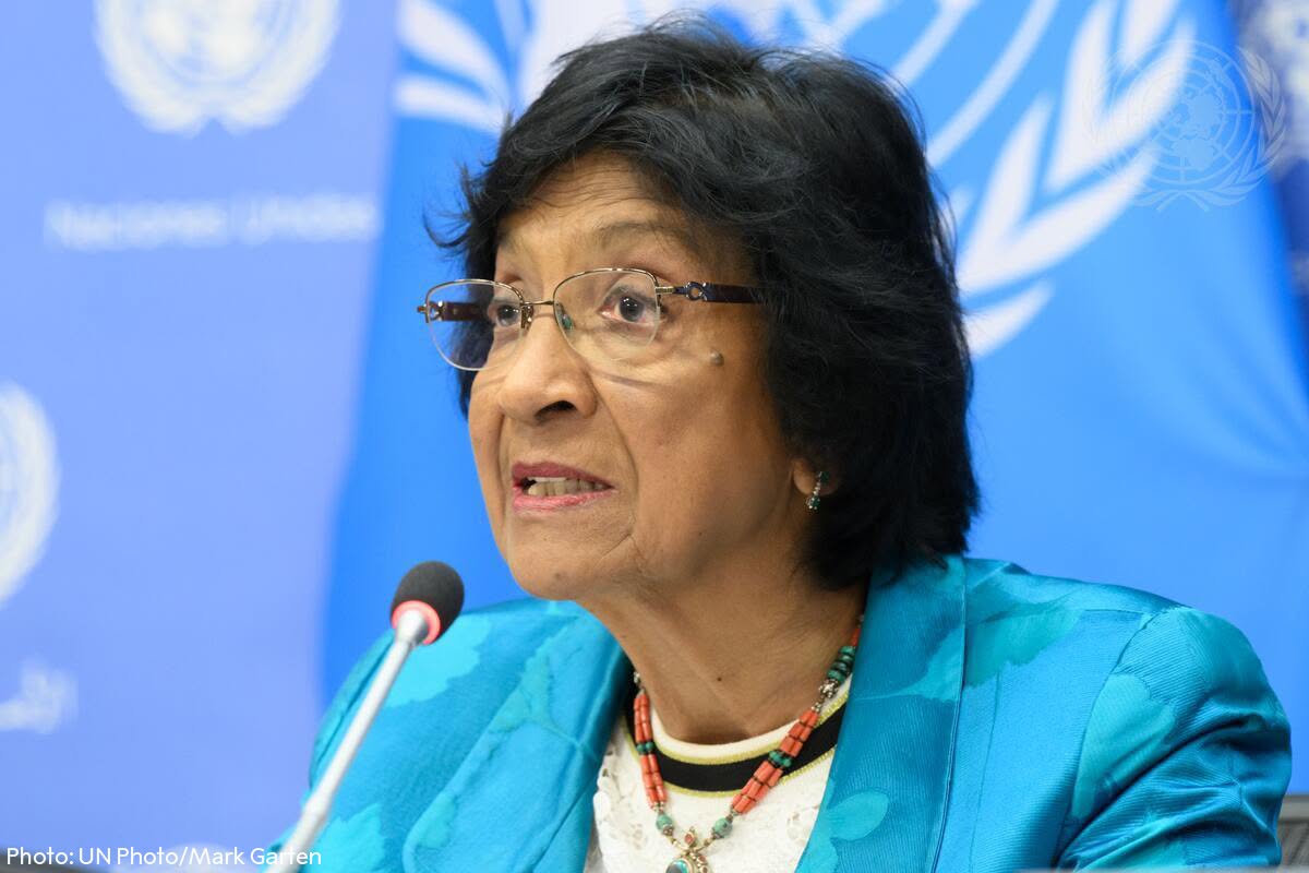 Woman speaking at a UN press briefing, wearing a turquoise blazer and beaded necklace, microphone in front.