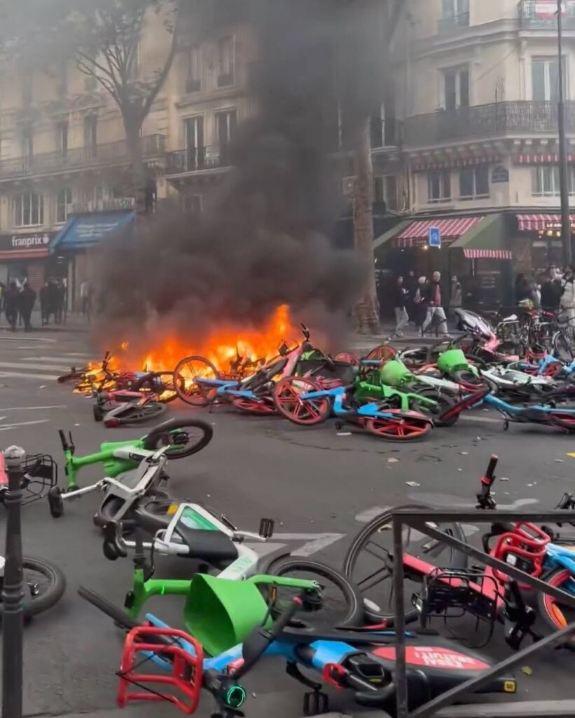 Bike rental bikes on a street piled and burning with thick black smoke rising into the air, flames visible among the wreckage in front of a building block.