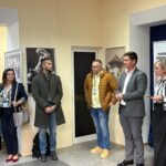 Group of people standing indoors along a wall with framed photos and a banner, as if at an event or briefing.