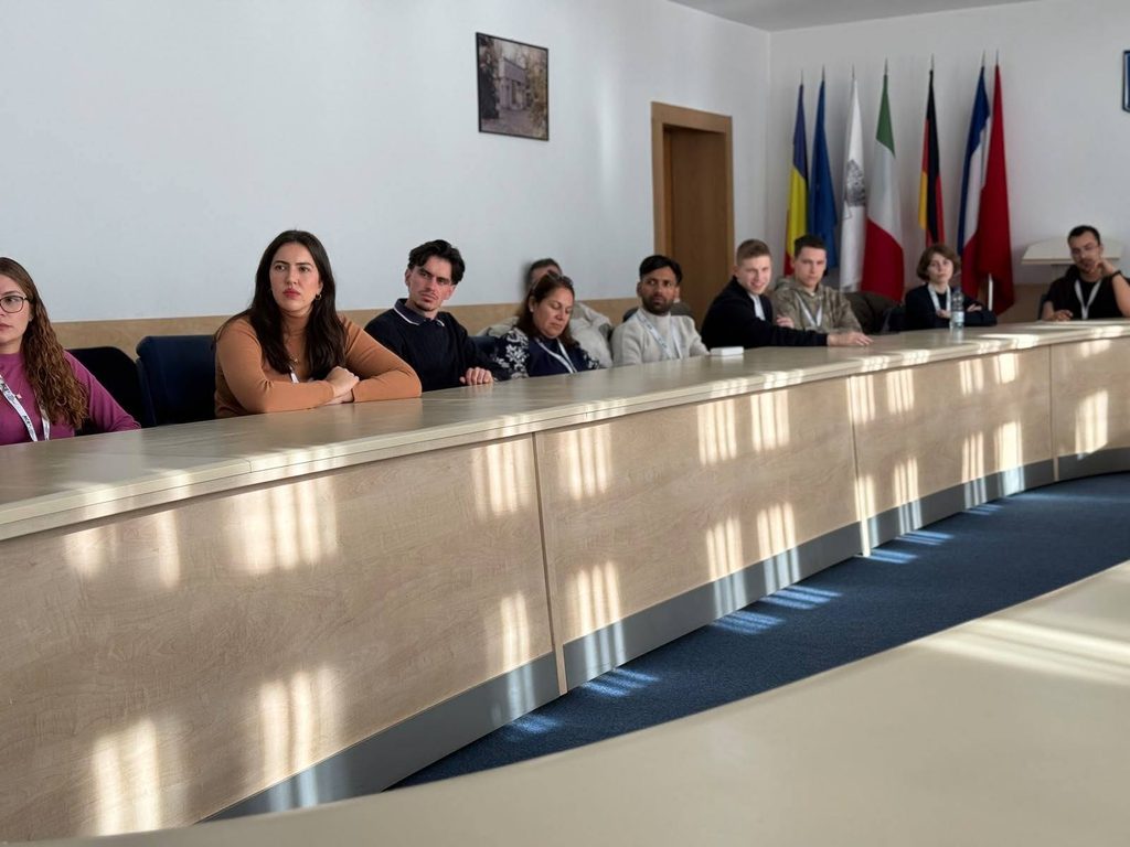 863772814_4615112455475385_38379464939169166_n - Jurnalul Olteniei – Știri din Craiova și Oltenia Group of people seated along a curved conference desk in a meeting room, with flags on the wall behind them.