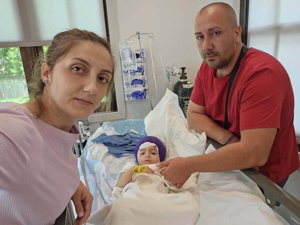 Woman in a pink top takes a selfie with a man in a red shirt beside a young patient in a hospital bed.