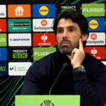 Man at a press conference podium, hand on cheek, with a wall of sponsor logos behind him.