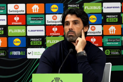 Man at a press conference podium, hand on cheek, with a wall of sponsor logos behind him.