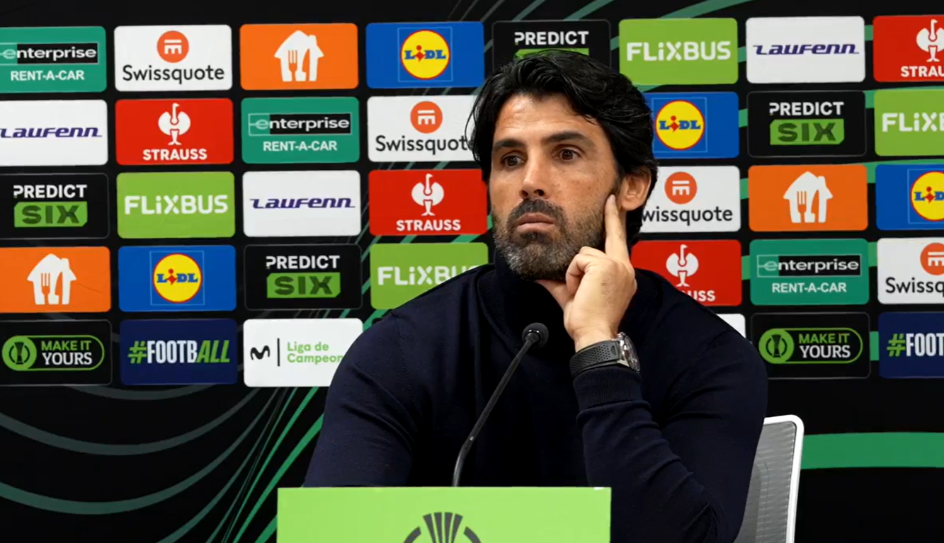 Man at a press conference podium, hand on cheek, with a wall of sponsor logos behind him.
