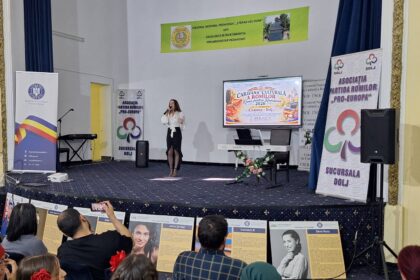 Female singer in a white blouse and black skirt performing on a stage with a large screen displaying an event banner behind her