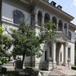 White historic villa with ornate arches, columns, and carved stonework, set among trees and a bench in the garden.