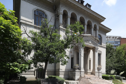 White historic villa with ornate arches, columns, and carved stonework, set among trees and a bench in the garden.