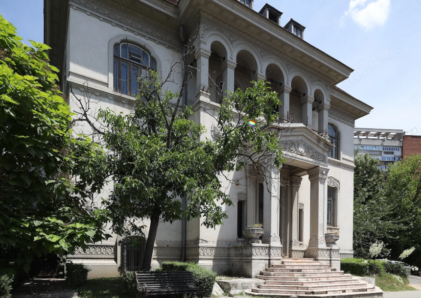 White historic villa with ornate arches, columns, and carved stonework, set among trees and a bench in the garden.