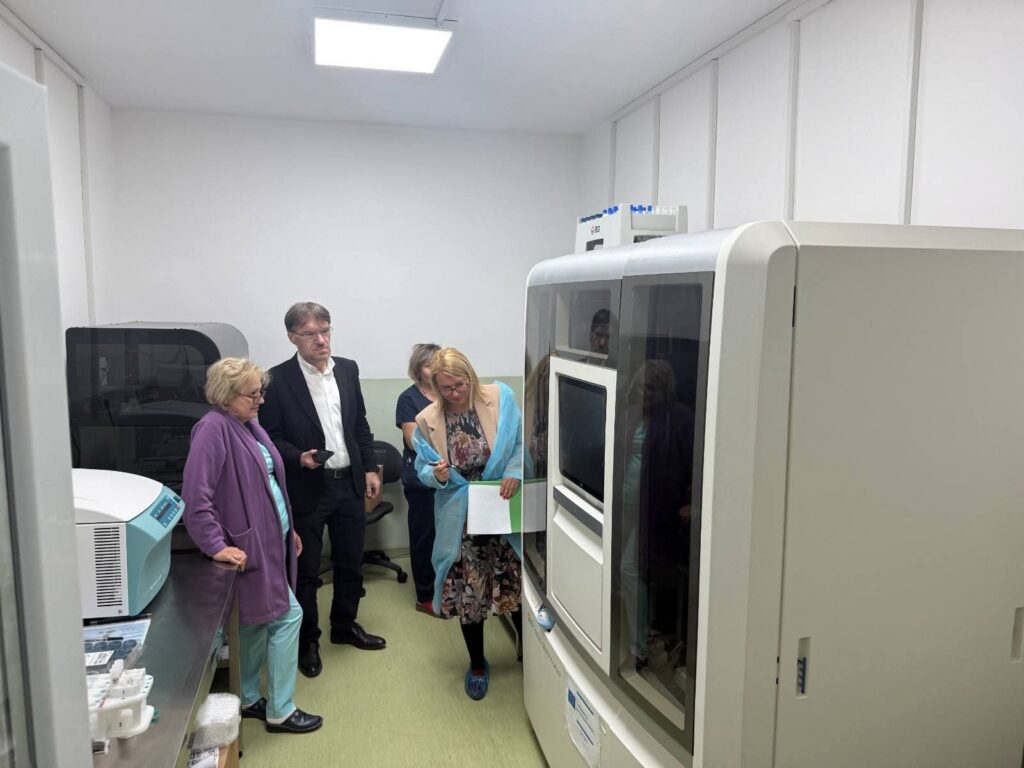 FB_IMG_1791349719299 - Jurnalul Olteniei – Știri din Craiova și Oltenia Four people in a clean lab observe a large automated lab instrument with a touchscreen.