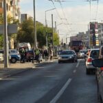 Busy city street scene with cars stopped at a crosswalk, police/emergency personnel nearby, and overhead tram lines crisscrossing the sky.