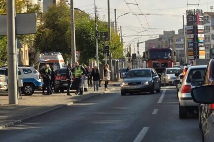 Busy city street scene with cars stopped at a crosswalk, police/emergency personnel nearby, and overhead tram lines crisscrossing the sky.