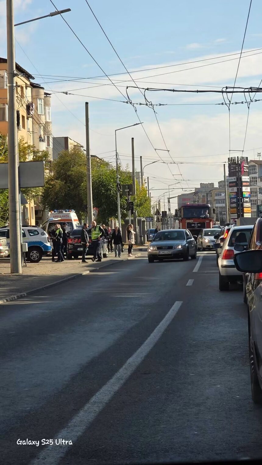 Busy city street scene with cars stopped at a crosswalk, police/emergency personnel nearby, and overhead tram lines crisscrossing the sky.