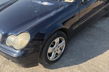 Black sedan with a cracked windshield and a damaged driver-side mirror in a lot, with police cars in the background