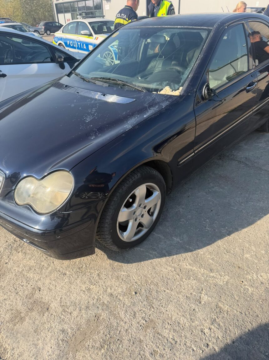 Black sedan with a cracked windshield and a damaged driver-side mirror in a lot, with police cars in the background