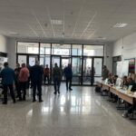 Crowd of people in a spacious lobby or hall with exhibitor tables along the sides and glass doors at the far end.