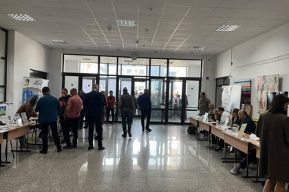 Crowd of people in a spacious lobby or hall with exhibitor tables along the sides and glass doors at the far end.