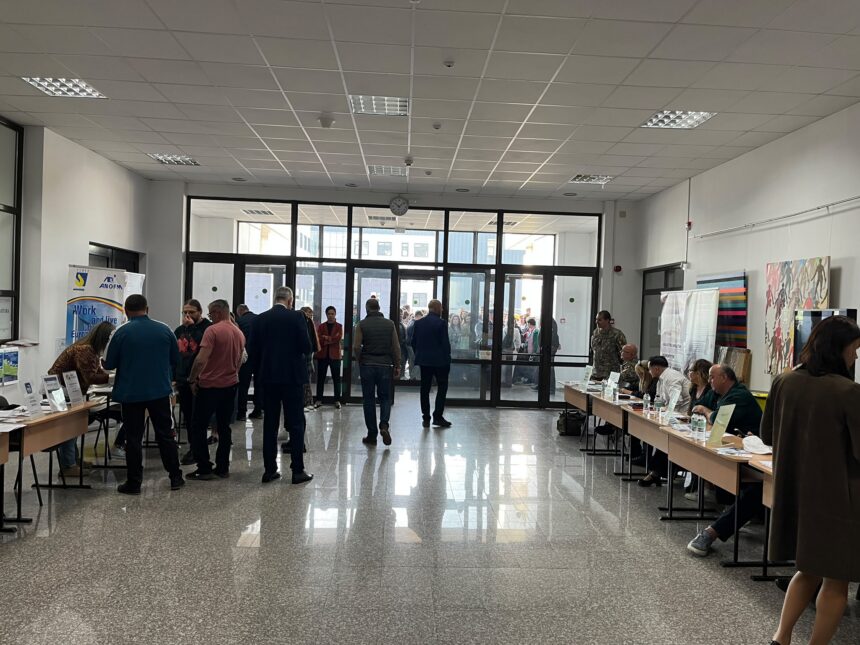 Crowd of people in a spacious lobby or hall with exhibitor tables along the sides and glass doors at the far end.