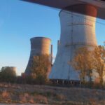 Row of tall cooling towers at an abandoned power plant, bathed in warm sunset light with trees in the foreground.