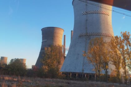 Row of tall cooling towers at an abandoned power plant, bathed in warm sunset light with trees in the foreground.