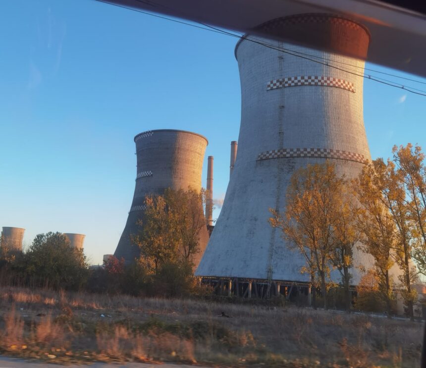 Row of tall cooling towers at an abandoned power plant, bathed in warm sunset light with trees in the foreground.