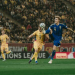 Two footballers—one in a blue Sweden kit (#4) and one in a yellow kit (#7)—jump to control the ball mid-air during a match, with a packed stadium in the background.