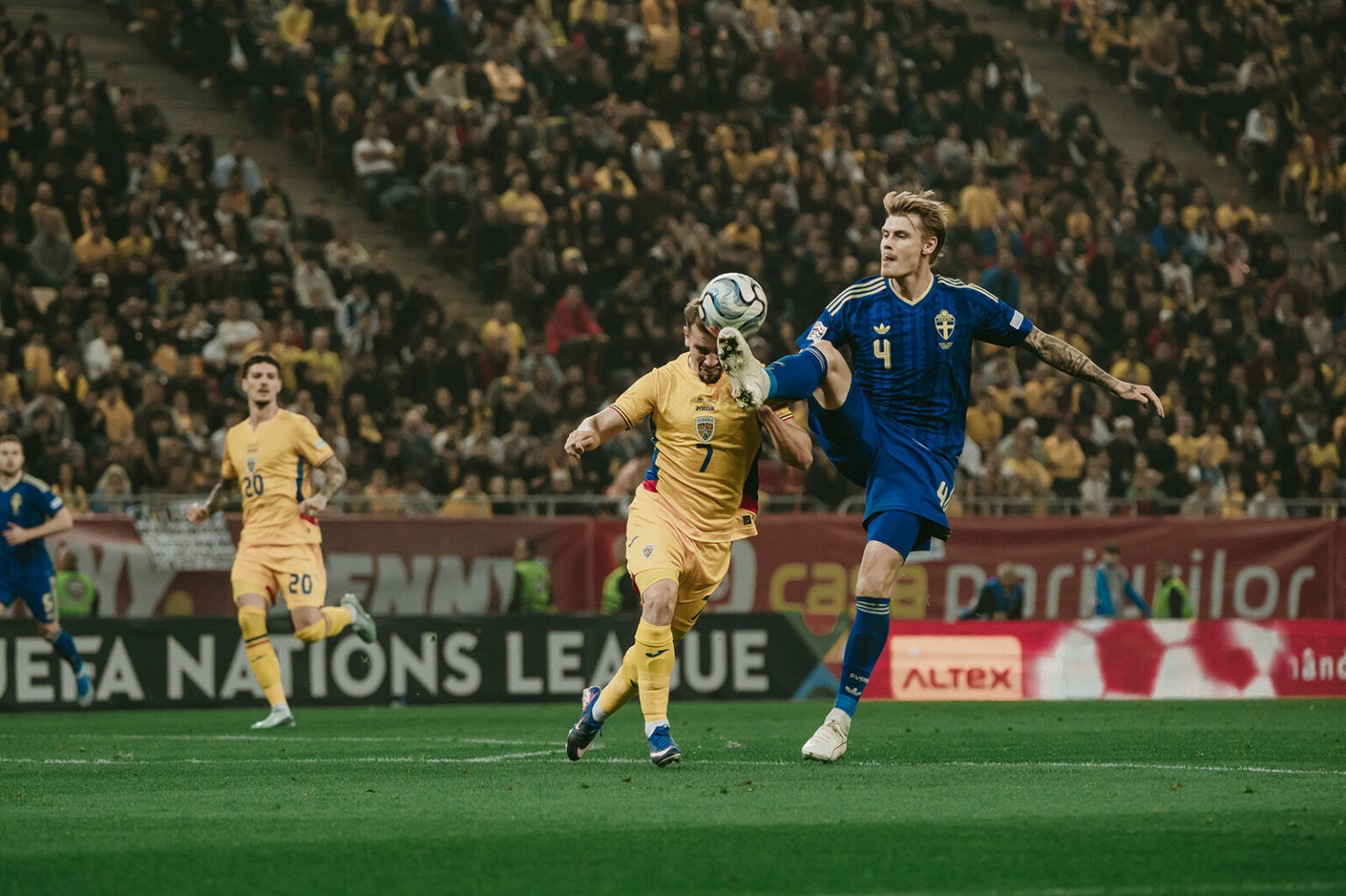 Two footballers—one in a blue Sweden kit (#4) and one in a yellow kit (#7)—jump to control the ball mid-air during a match, with a packed stadium in the background.
