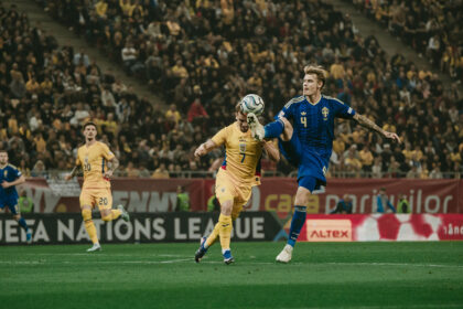 Two footballers—one in a blue Sweden kit (#4) and one in a yellow kit (#7)—jump to control the ball mid-air during a match, with a packed stadium in the background.