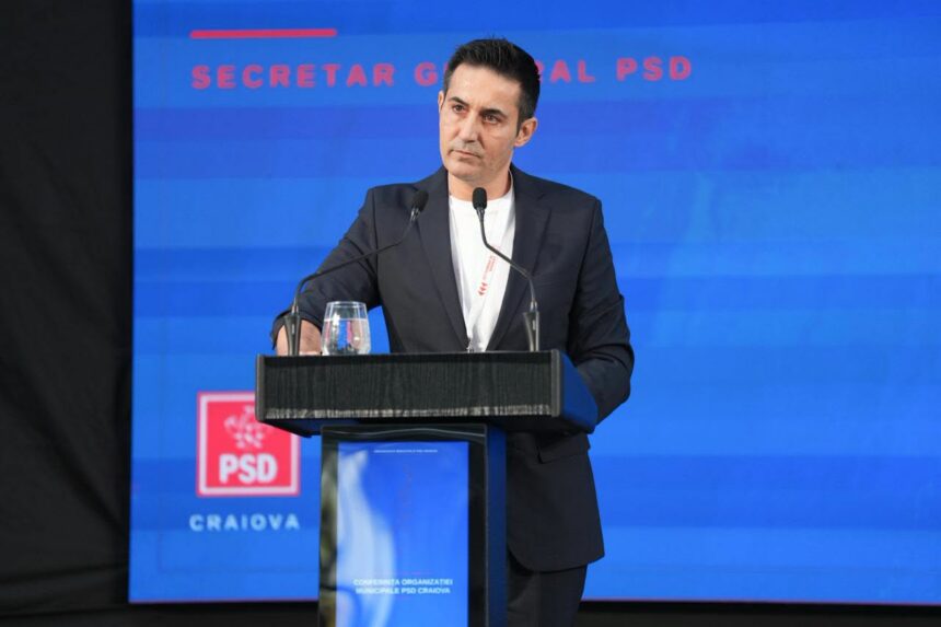 Man in a dark suit standing at a podium with two microphones, delivering a speech against a blue PSD banner backdrop.