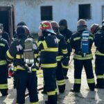 Group of firefighters in turnout gear with air tanks standing in a circle outside a building, likely during a drill or briefing.