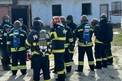 Group of firefighters in turnout gear with air tanks standing in a circle outside a building, likely during a drill or briefing.
