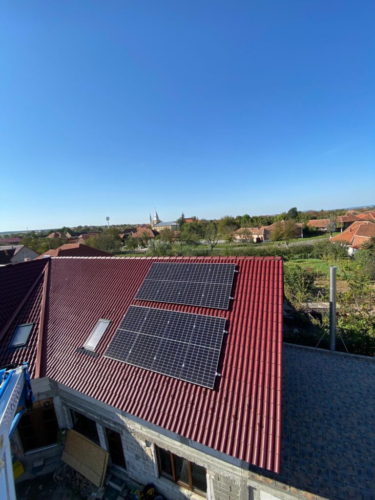 Residential rooftop with solar panels mounted on a red tiled roof against a clear blue sky; distant village with church spires visible on the horizon.