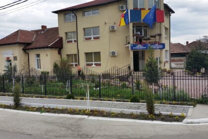 Two-story beige government building with blue and yellow flags, a fenced garden, and a paved entrance area nearby.