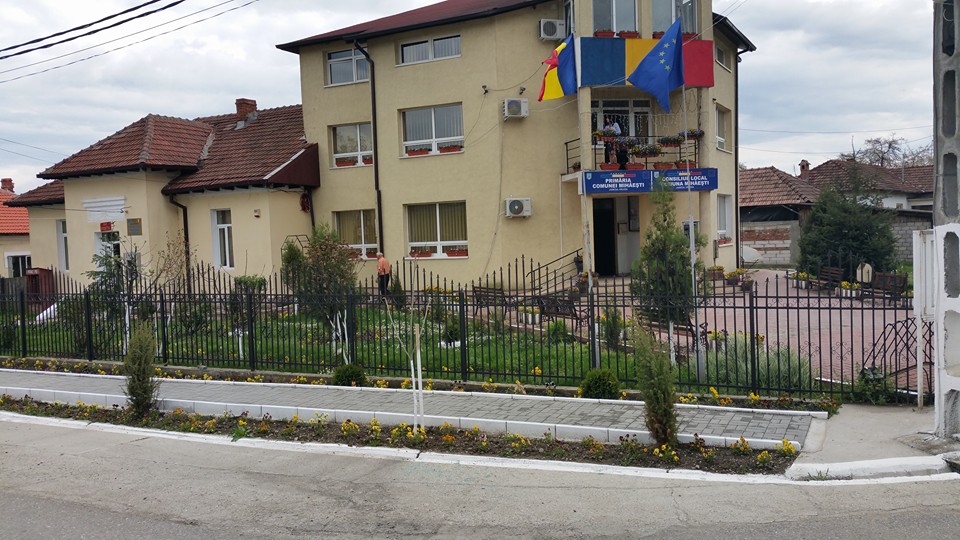 Two-story beige government building with blue and yellow flags, a fenced garden, and a paved entrance area nearby.