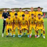 Youth soccer team in yellow uniforms posing for a group photo on a grassy field, with a goalkeeper in black on the left.