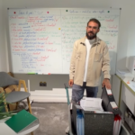 Instructor standing beside a whiteboard filled with colorful notes, in a small classroom with a filing cart and chair nearby.