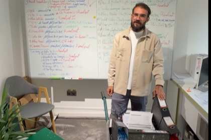Instructor standing beside a whiteboard filled with colorful notes, in a small classroom with a filing cart and chair nearby.