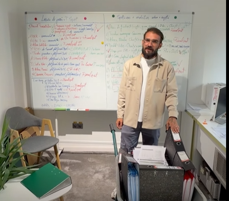 Instructor standing beside a whiteboard filled with colorful notes, in a small classroom with a filing cart and chair nearby.
