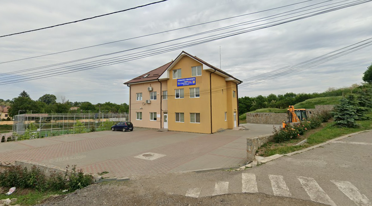 primaria mihaesti olt - Jurnalul Olteniei – Știri din Craiova și Oltenia Yellow three-story building with a blue sign, a small car parked at the entrance, and a construction excavator on the right.