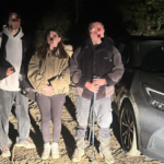 Three people with backpacks pose on a dirt road at night beside a gray SUV; faces blurred for privacy.