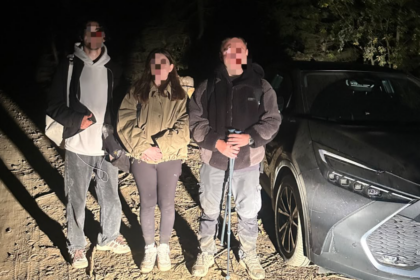 Three people with backpacks pose on a dirt road at night beside a gray SUV; faces blurred for privacy.