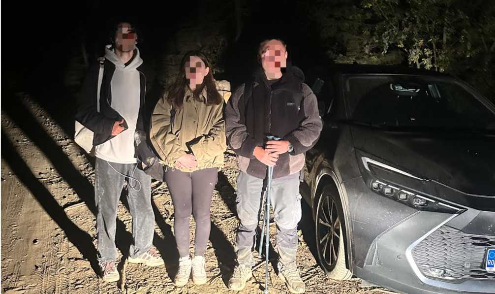 Three people with backpacks pose on a dirt road at night beside a gray SUV; faces blurred for privacy.