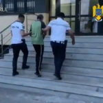 Three plainclothes officers escort a man up the building stairs outside a government facility, with police logos visible at top corners (Romania).