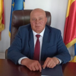 Bald man in a blue suit and red tie sits at a wooden desk in an office, with flags behind him and hands resting on the desk.